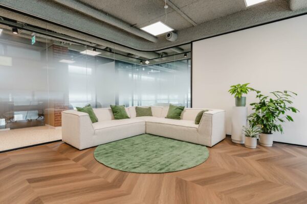 Moderne loungehoek met witte hoekbank, groene kussens, planten en een groen vloerkleed in een kantoor aan de Joop Geesinkweg.